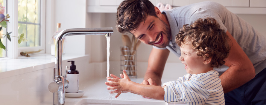 Un padre y un niño pequeño se ríen lavándose las manos en un lavabo blanco con un grifo plateado. Un dispensador de jabón está al lado.