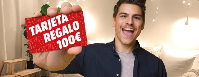 Hombre sonriente sujetando una tarjeta regalo roja con texto blanco "TARJETA REGALO 100€". Fondo con cortinas y luces decorativas.