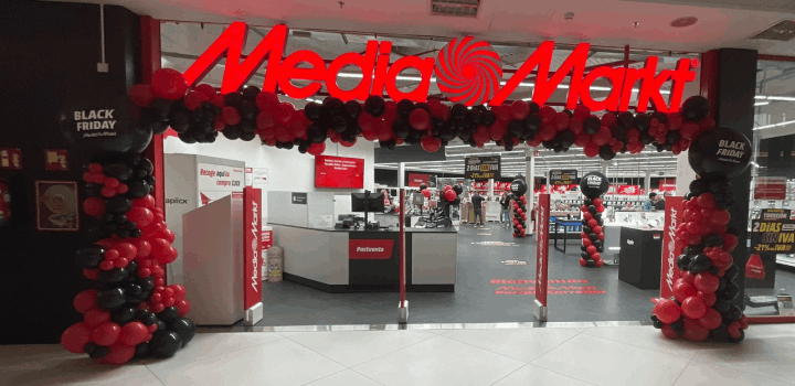 Entrada a Media Markt decorada con letrero rojo, globos rojos y negros. Dentro, cajas registradoras y pasillos con productos.