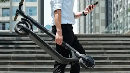 Un hombre de camisa blanca sostiene un patinete plegado y un móvil en unas escaleras grises, con edificios al fondo.