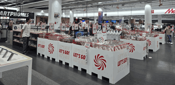 Interior de una tienda con mostradores blancos que anuncian "Let's Go" y un espiral rojo. Gente y estantes con productos alrededor.