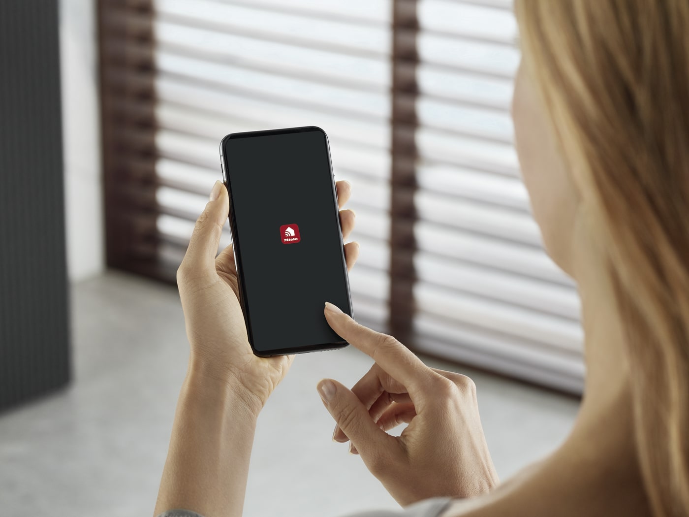 Mujer usa móvil con logo rojo sobre pantalla negra. Persianas horizontales al fondo.