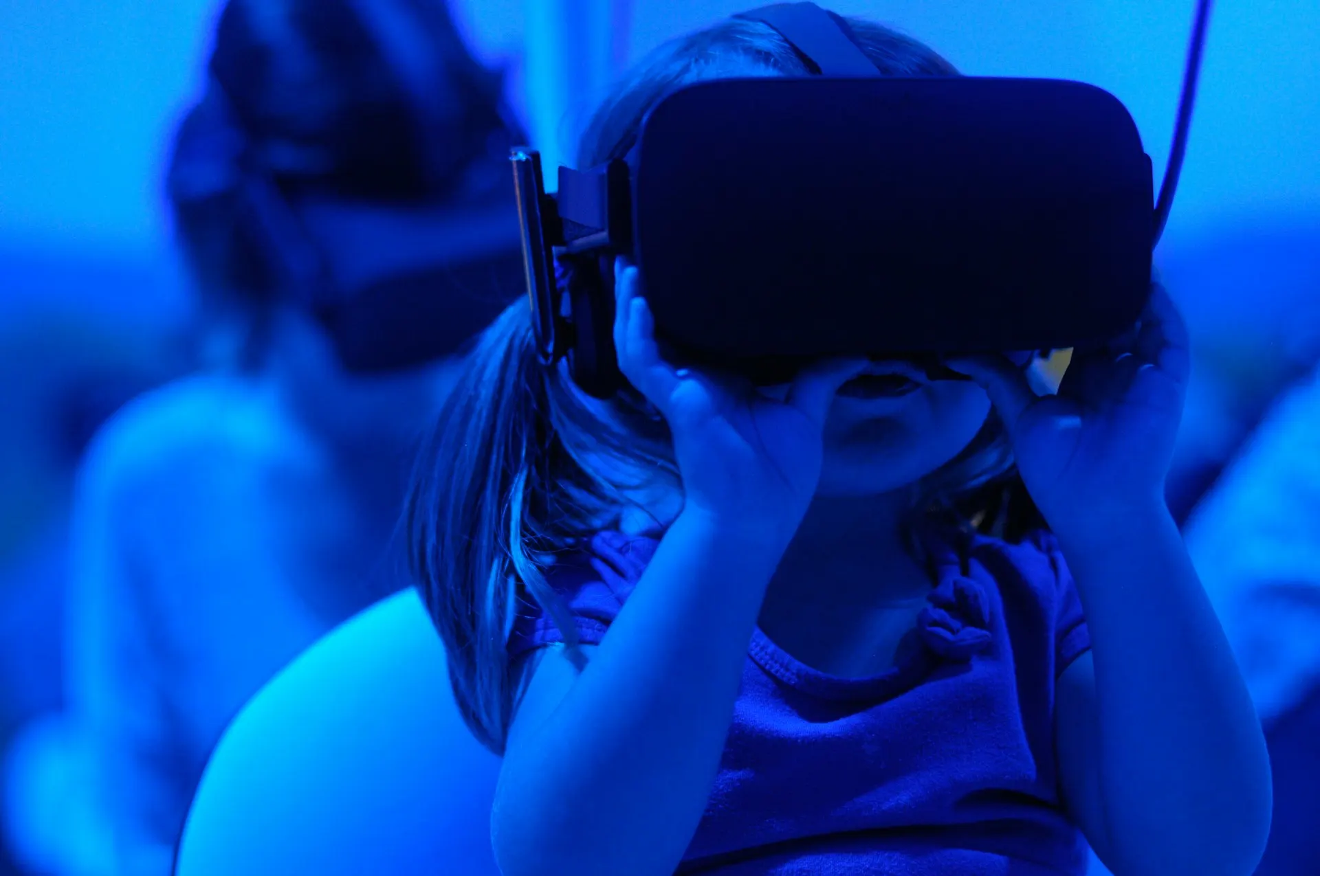 Niña con camiseta azul y gafas de realidad virtual, iluminada en azul, con otra persona borrosa al fondo.