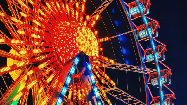 Una noria iluminada con luces naranjas, amarillas y azules contra un cielo nocturno oscuro. La estructura es de metal con cabinas rojas.