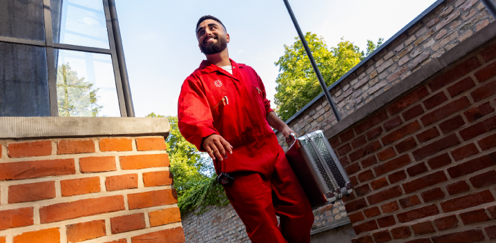 Un hombre con un mono rojo lleva un maletín plateado junto a una pared de ladrillo. El cielo es azul y hay árboles al fondo.