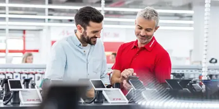 Bir mağazada, sakallı bir müşteri ve kırmızı gömlekli bir çalışan tabletleri inceliyorlar. Raflarda ürünler var.