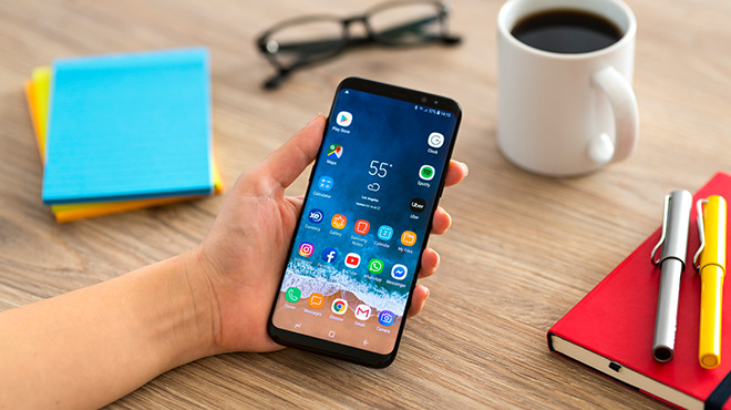 Bir el, bir akıllı telefonu tutuyor; ekranda hava durumu uygulaması açık. Masada not defteri, kalemler, kahve ve gözlükler var.