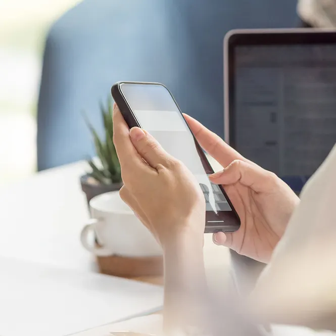 Elleriyle telefon kullanan kişi. Arka planda dizüstü bilgisayar ve bitki görülüyor.