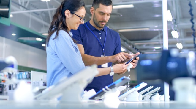 Müşteri ve mağaza görevlisi bir elektronik mağazasında bir telefonu inceliyor.