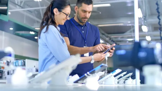 Müşteri ve mağaza görevlisi bir elektronik mağazasında bir telefonu inceliyor.