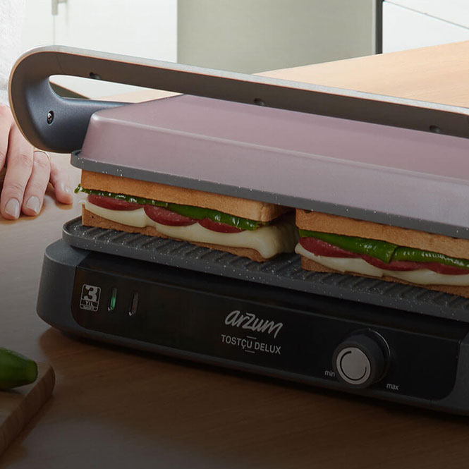 Arzum tost makinesi, iki adet sandviçi pişiriyor. Sandviçler peynir, domates ve yeşillik içeriyor.