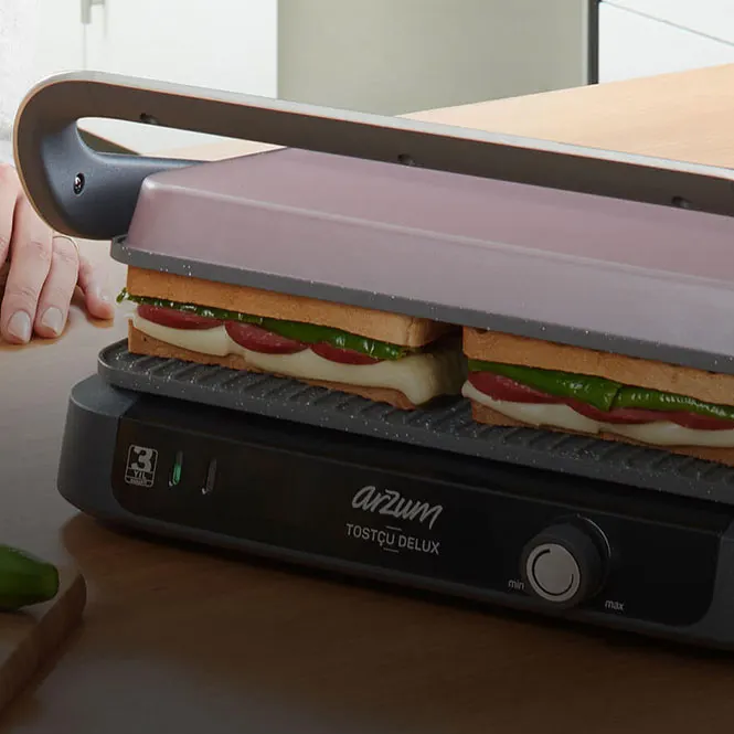 Arzum tost makinesi, iki adet sandviçi pişiriyor. Sandviçler peynir, domates ve yeşillik içeriyor.