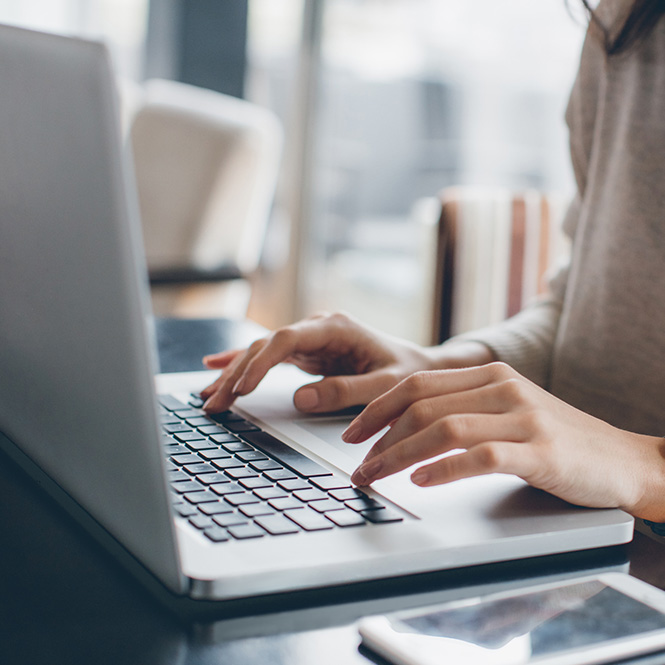 Bir kadın, gri bir dizüstü bilgisayarda yazı yazıyor. Beyaz bir telefon masanın üzerinde duruyor. Arka planda bulanık bir pencere var.