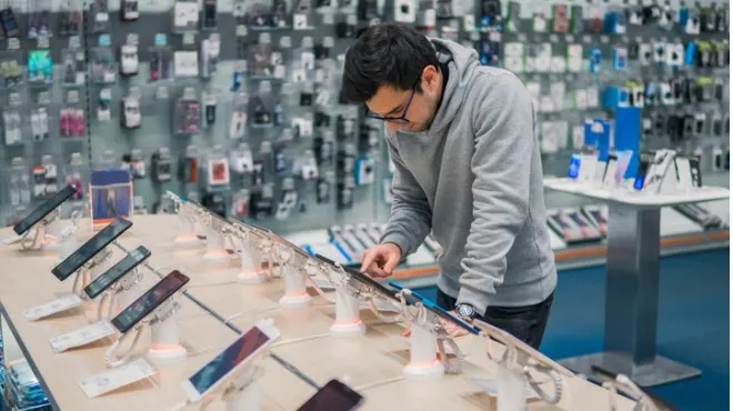 Cep telefonu mağazasında telefonları inceleyen gri kapüşonlu bir adam. Raflarda çok sayıda telefon aksesuarı var.