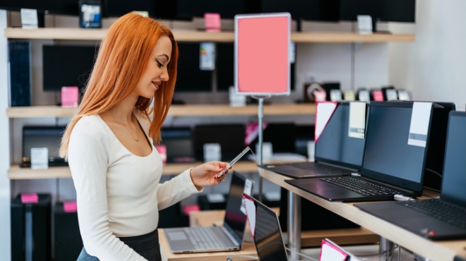 Kırmızı saçlı kadın, elektronik mağazasında dizüstü bilgisayarlara bakıyor. Raflarda pembe etiketler ve ekranlar var.