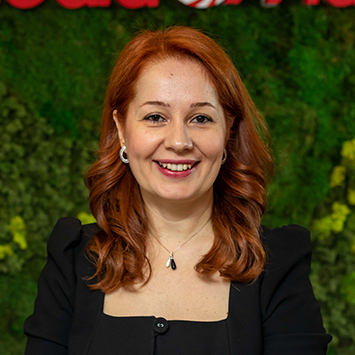 Kızıl saçlı, gülen kadın, siyah ceketli, yeşil bitki örtüsü önünde, kolye ve küpe takıyor.