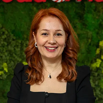 Kızıl saçlı, gülen kadın, siyah ceketli, yeşil bitki örtüsü önünde, kolye ve küpe takıyor.