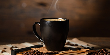 Siyah fincanda kahve, ahşap tepside kahve çekirdekleri arasında duruyor. Buhar yükseliyor.