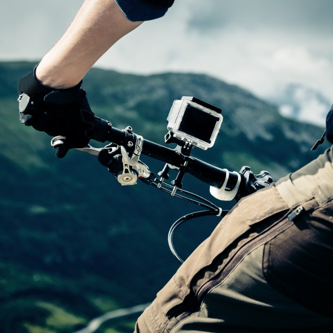 Dağ bisikletinin gidonunda aksiyon kamerası tutan bir bisikletçi. Arka planda dağlar ve gökyüzü var.