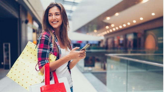 Alışveriş merkezinde cep telefonu kullanan gülümseyen kadın. Çantalar taşıyor. Arka planda mağazalar var.