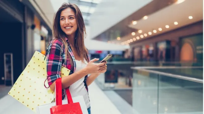 Alışveriş merkezinde cep telefonu kullanan gülümseyen kadın. Çantalar taşıyor. Arka planda mağazalar var.