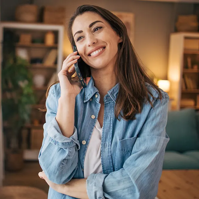 Gülümseyen kadın, mavi gömlekli, cep telefonuyla konuşuyor. Arka planda kitaplık ve bitkiler var.