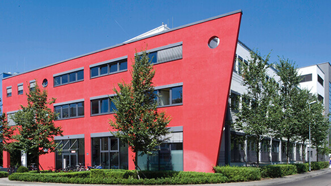 Kırmızı ve gri renklerde, çok katlı, modern bir bina ve önünde ağaçlar ile çalılar bulunuyor.