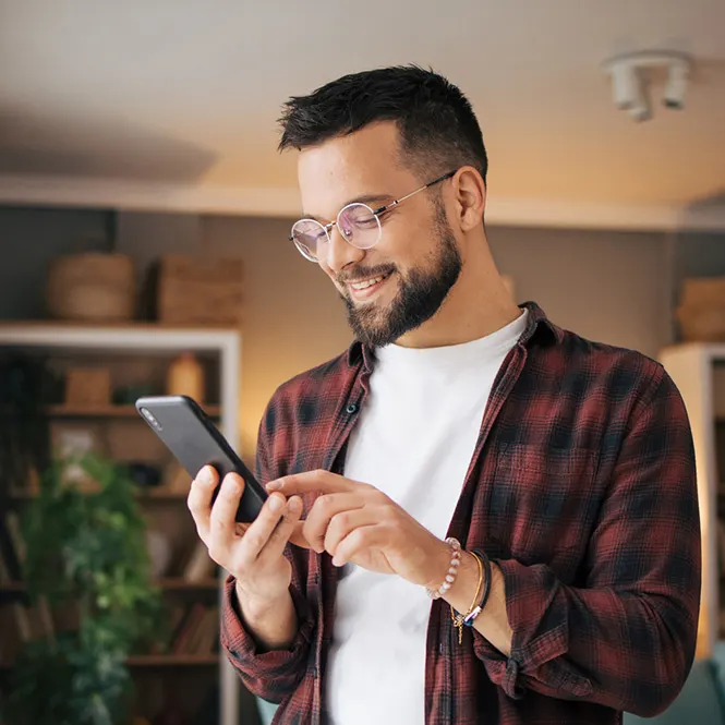 Sakallı, gözlüklü bir adam, kırmızı ekose gömlek ve beyaz tişört giyiyor, elinde cep telefonu var ve gülümsüyor.