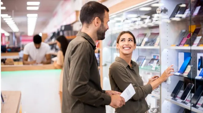 Bir mağazada, bir kadın ve bir adam cep telefonlarını inceliyor. Kadın gülümserken, adam elinde bir not tutuyor.