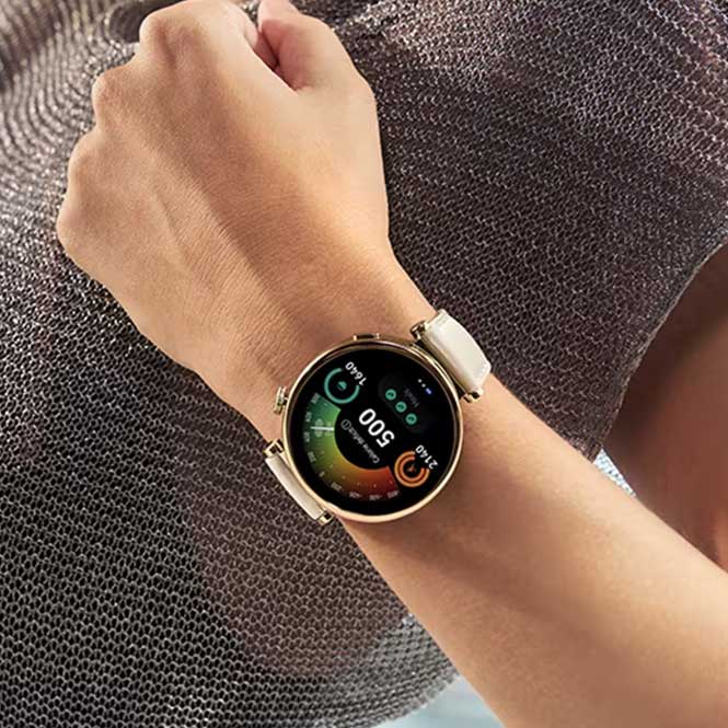 Kolda yuvarlak, altın rengi akıllı saat var. Ekranda 500 sayısı, renkli grafikler ve simgeler görülüyor. Arka planda örgü desenli gri bir kumaş var.