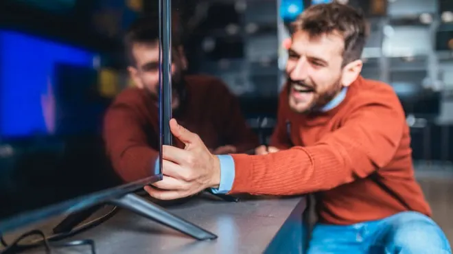 Gülümseyen bir adam, elektronik mağazasında yeni bir TV'yi inceliyor; turuncu bir kazak ve mavi kot pantolon giyiyor.