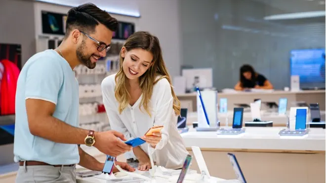 Bir mağazada cep telefonlarına bakan bir adam ve bir kadın. Tezgahta sergilenen telefonlar ve arka planda bir çalışan görülüyor.
