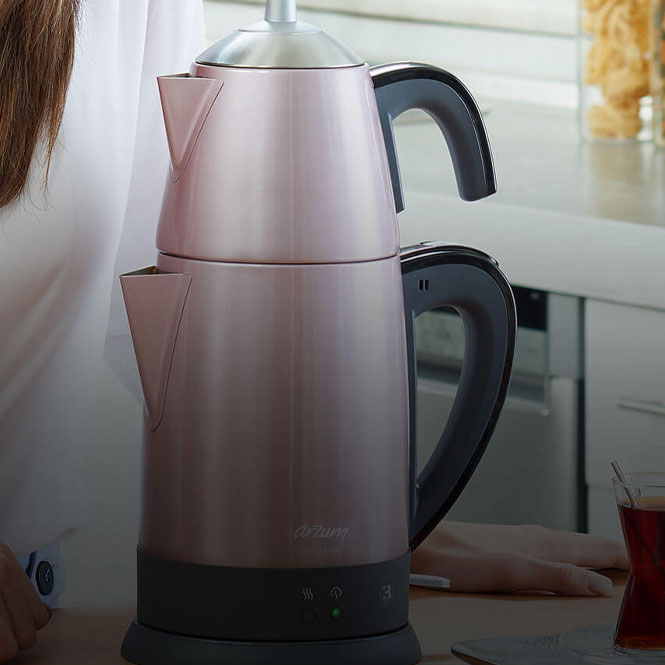 Pembe çay makinesi, siyah detaylar, tezgah üzerinde. Arka planda mutfak ve kısmen görünen bir kişi yer alıyor.