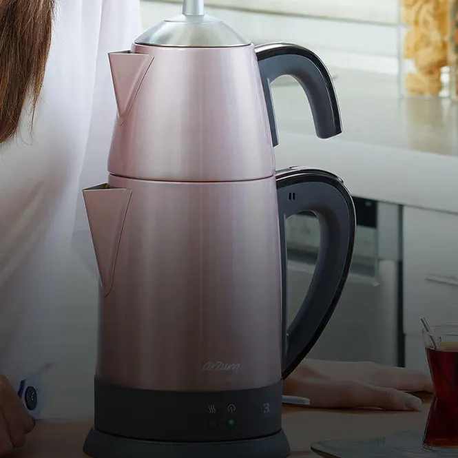 Pembe çay makinesi, siyah detaylar, tezgah üzerinde. Arka planda mutfak ve kısmen görünen bir kişi yer alıyor.