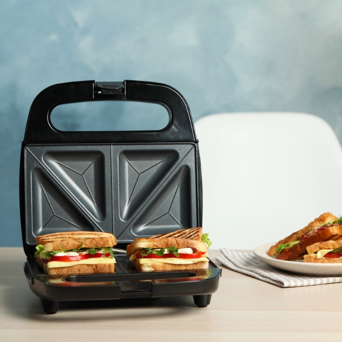 Siyah tost makinesi, iki sandviç ve tabakta tostlar. Arka planda mavi duvar ve beyaz sandalye var.