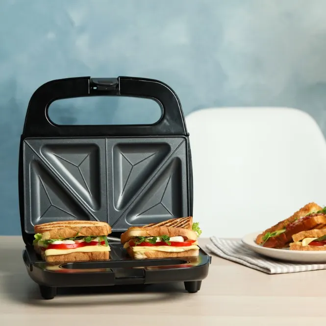 Siyah tost makinesi, iki sandviç ve tabakta tostlar. Arka planda mavi duvar ve beyaz sandalye var.