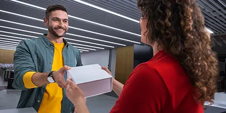 Bir mağazada bir kadın beyaz bir kutuyu kırmızı tişörtlü bir adamdan alıyor. Kadın gülümsüyor ve ekose bir gömlek giyiyor.