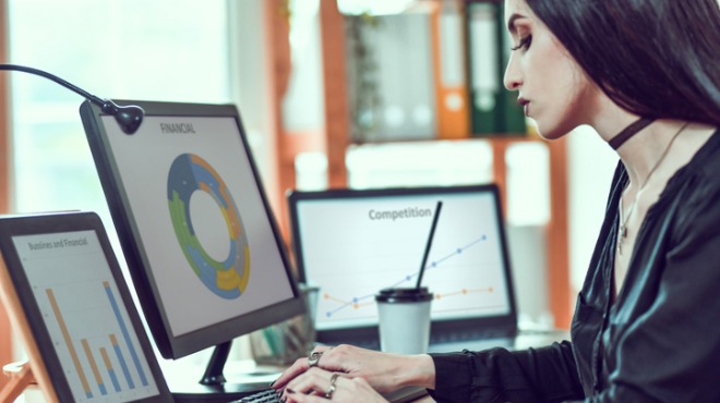 Siyah saçlı kadın, ofiste bilgisayar başında çalışıyor. Ekranda grafikler ve kahve bardağı görülüyor.