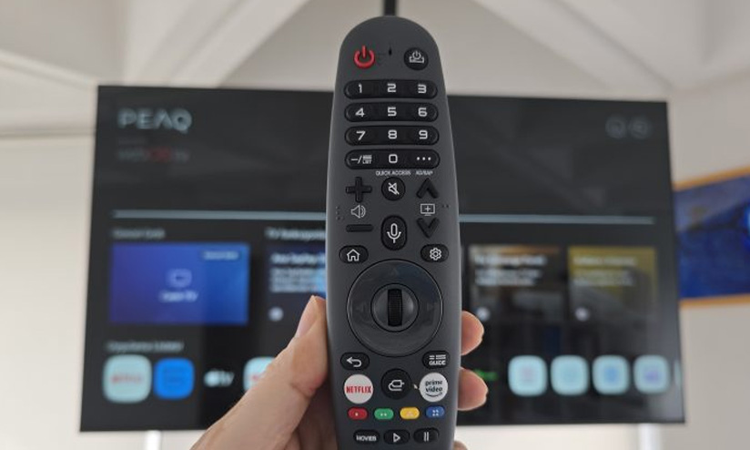 Bir elin tuttuğu, düğmeleri olan siyah bir uzaktan kumanda ve arka planda bir televizyon görülmektedir.