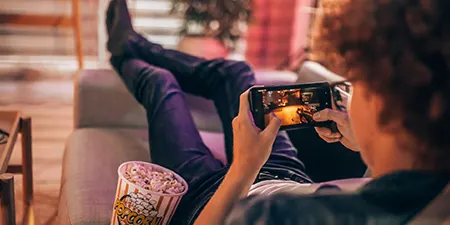 Koltukta uzanmış, telefonla oyun oynayan, kucağında patlamış mısır olan bir kişi. Arka planda iç mekan ve mobilyalar görülüyor.