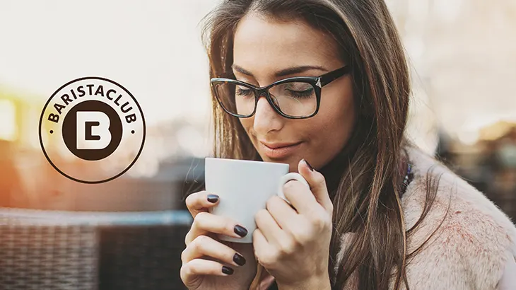 Kahverengi saçlı kadın, gözlük takmış, beyaz fincandan içiyor. Sol üstte "BaristaClub" logosu var.