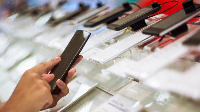 Bir el, siyah bir akıllı telefonu tutuyor. Arka planda, sergilenen diğer telefonlar ve aksesuarlar bulanık bir şekilde görülüyor.