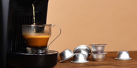 Kahverengi fonda kahve makinesi, kahve dolu fincan ve alüminyum kahve kapsülleri görülmektedir.