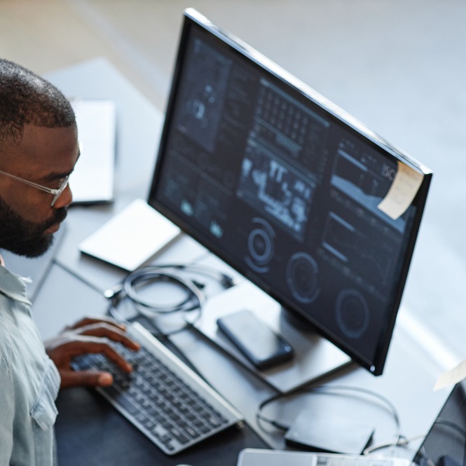 Siyah sakallı bir adam, karmaşık grafiklerle dolu bir bilgisayar ekranına bakarak klavyede yazı yazıyor.