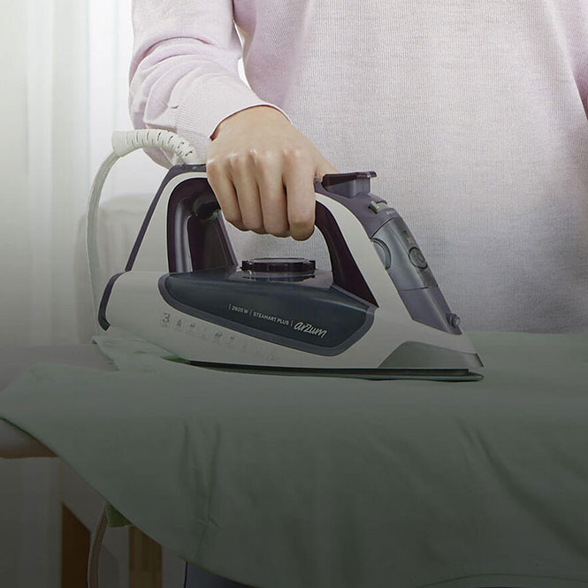 Kişi, ütü masasında Arzum marka, gri ve beyaz renkli bir ütüyü tutuyor. Kişi pembe bir kazak giyiyor.