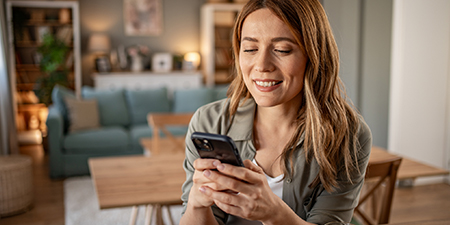 Gülümseyen bir kadın, oturma odasında telefon kullanıyor. Arka planda kanepe ve masa bulunuyor.