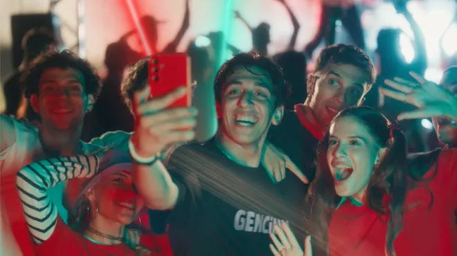 Kalabalık bir partide, bir grup genç kırmızı bir telefonla selfie çekiyor, gülümsüyor ve eğleniyor.