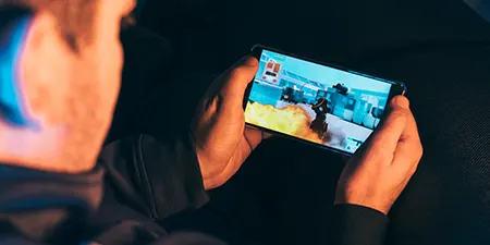 Bir kişi, elleriyle yatay olarak tuttuğu bir telefonda oyun oynuyor. Telefonda aksiyon dolu bir oyun sahnesi görülüyor.