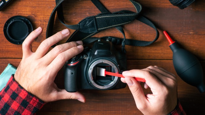 Ahşap zeminde, bir kişi kırmızı saplı fırça ile siyah kameranın içini temizliyor. Yanında temizleme araçları var.