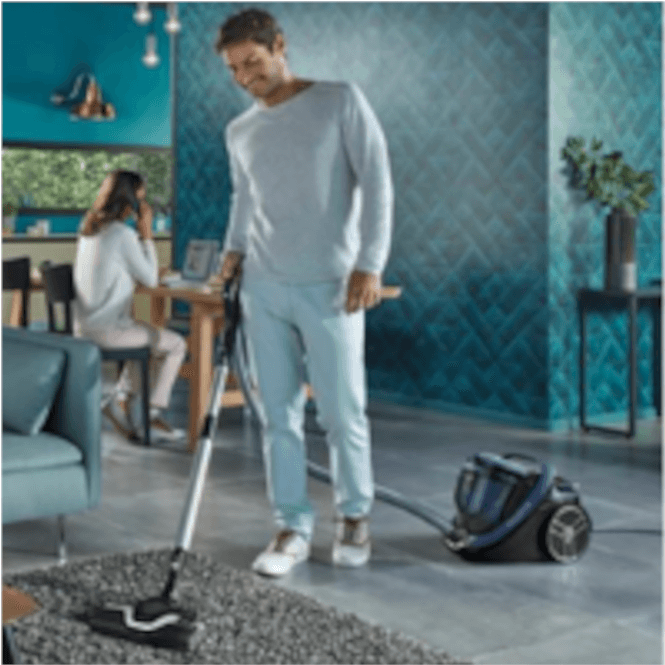 Adam halı üzerinde elektrikli süpürgeyle temizlik yapıyor. Arka planda kadın oturuyor.